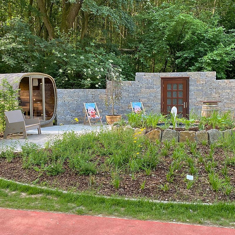 Ein Garten mit einer Sauna umrandet von Steinen und im Hintergrund ist ein Wald