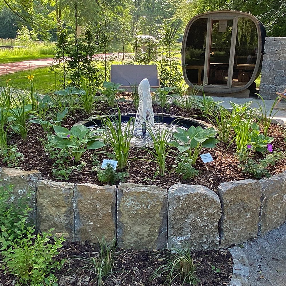 Ein runder Steingarten mit Bepflanzung und einer Außensauna im Hintergrund